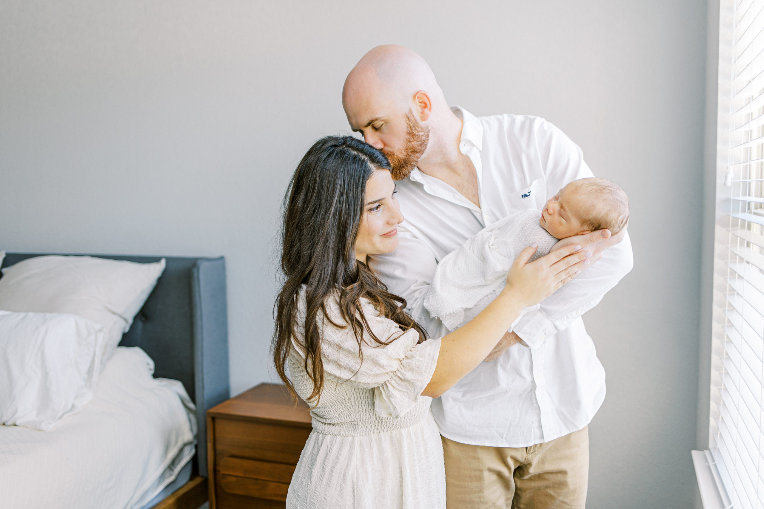 Husband leans over to kiss wife while holding their newborn son during lifestyle newborn session with newborn photographer dallas ling waters photography
