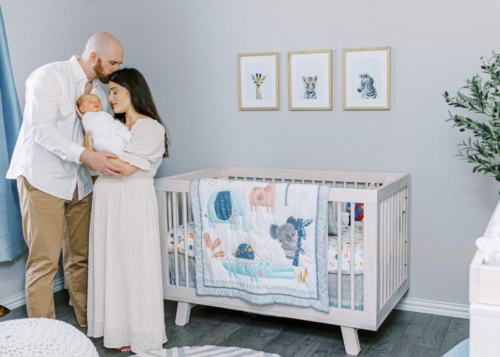 dad faces mom and closes his eyes as he kisses her on the forehead while she closes her eyes and holds newborn baby next to crib in nursery during a mckinney newborn photography with ling waters photography
